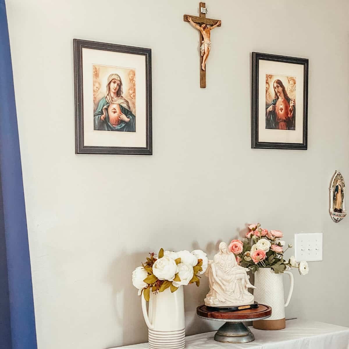 home altar with crucifix and Sacred & Immaculate heart frames above, two pitchers of white and pink flowers on each side