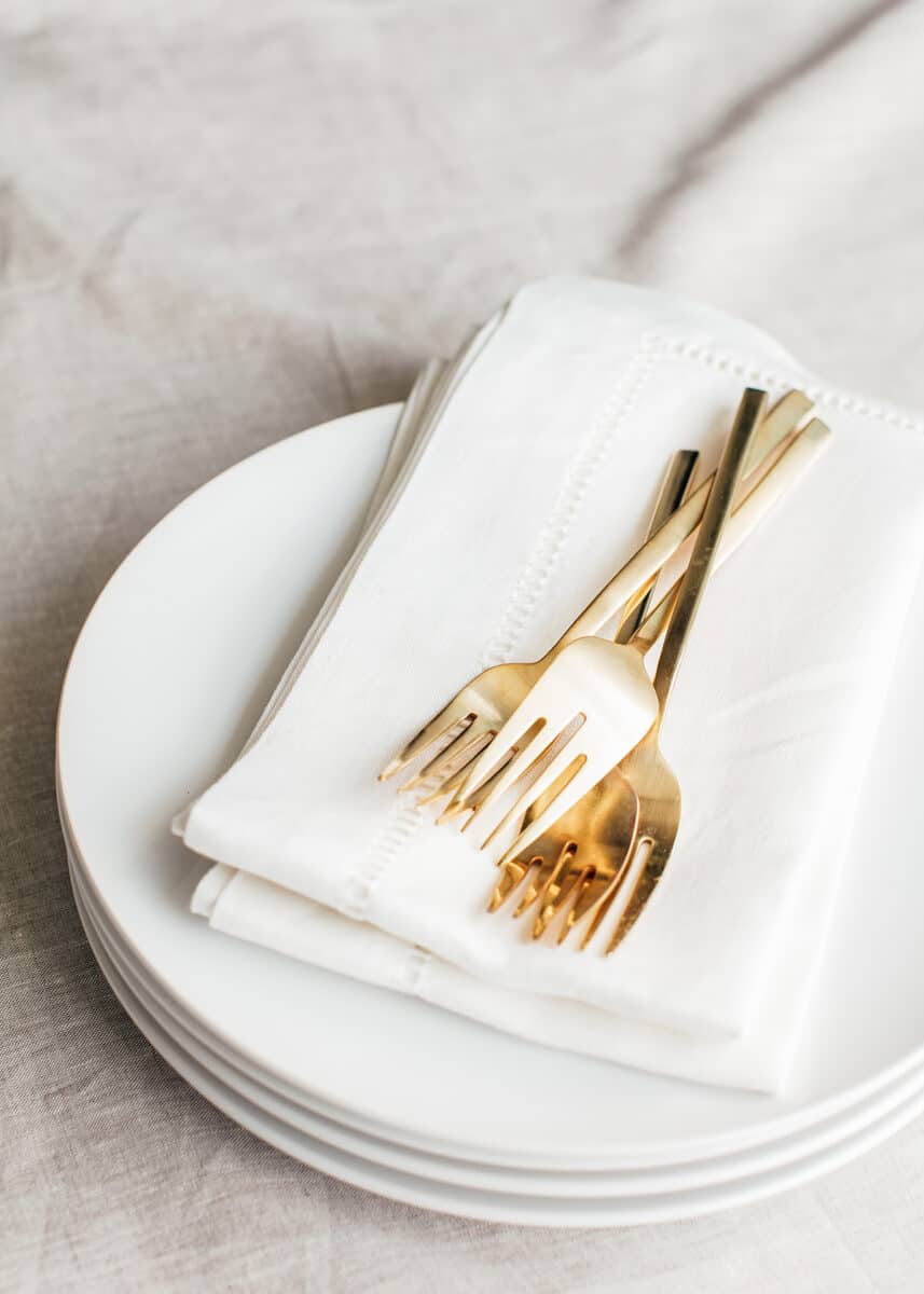 white linen napkins folded on a stack of white plates with gold forks.