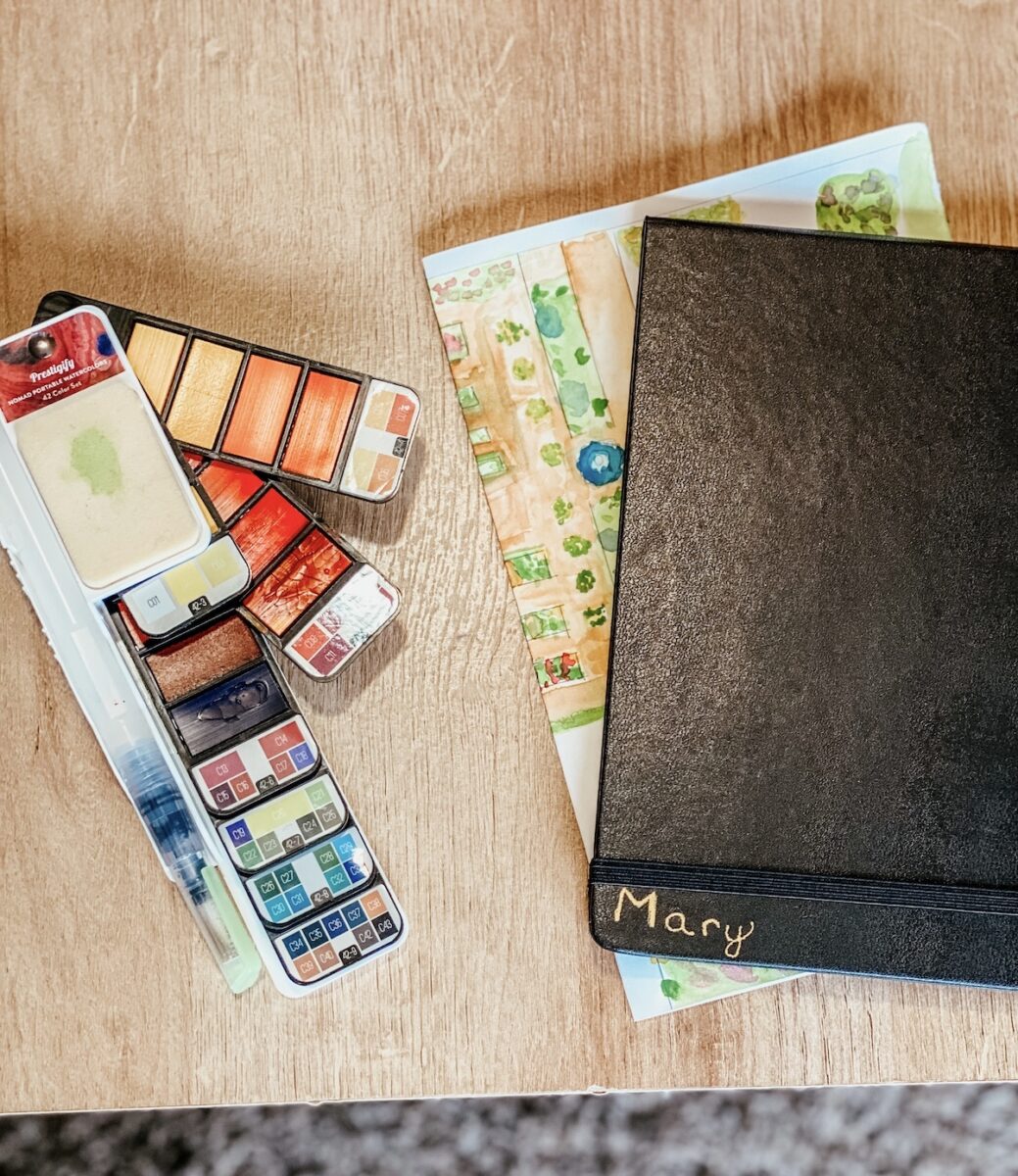 nature journal, watercolor pallet, and sketch of garden on a table.