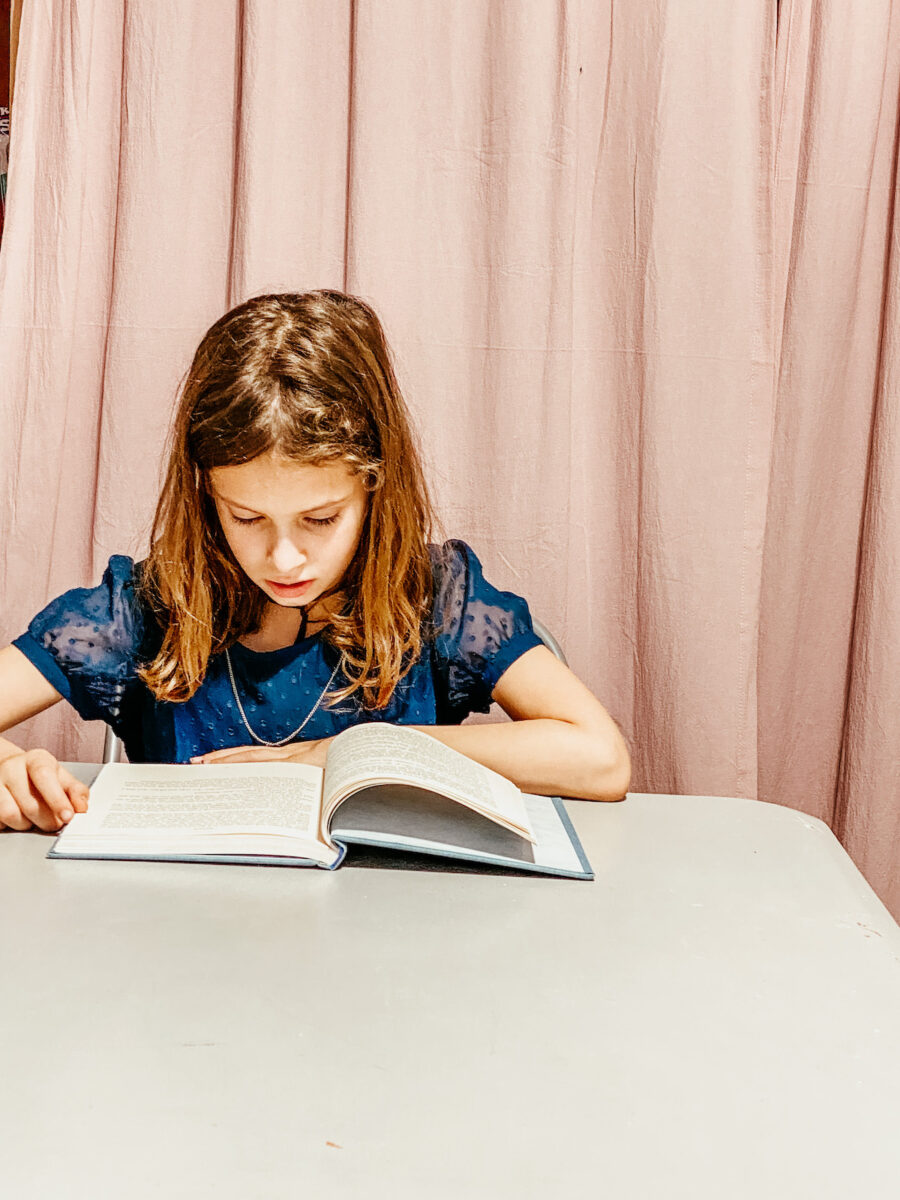 little girl sitting at a table reading a novel.
